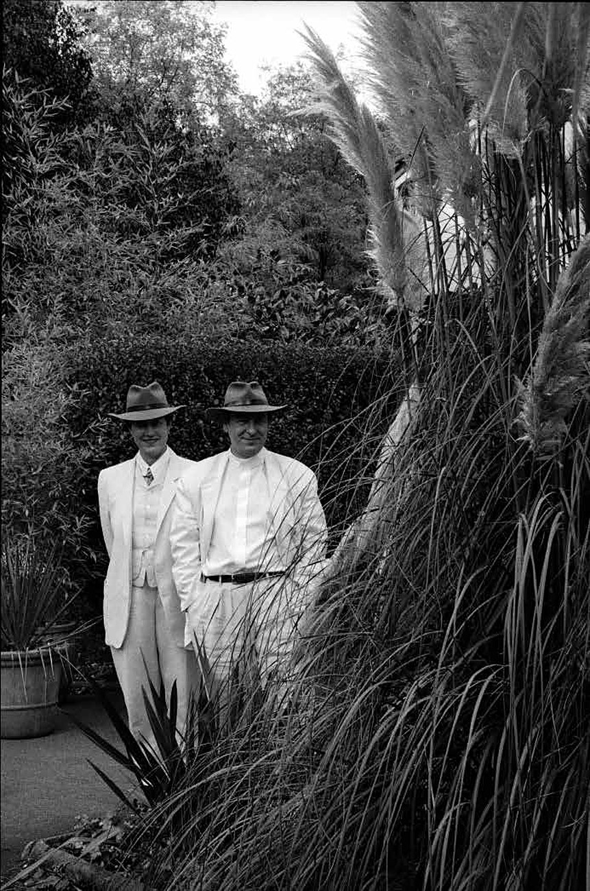 Black-and-white photograph: two figures dressed in white, wearing hats, in a lush garden